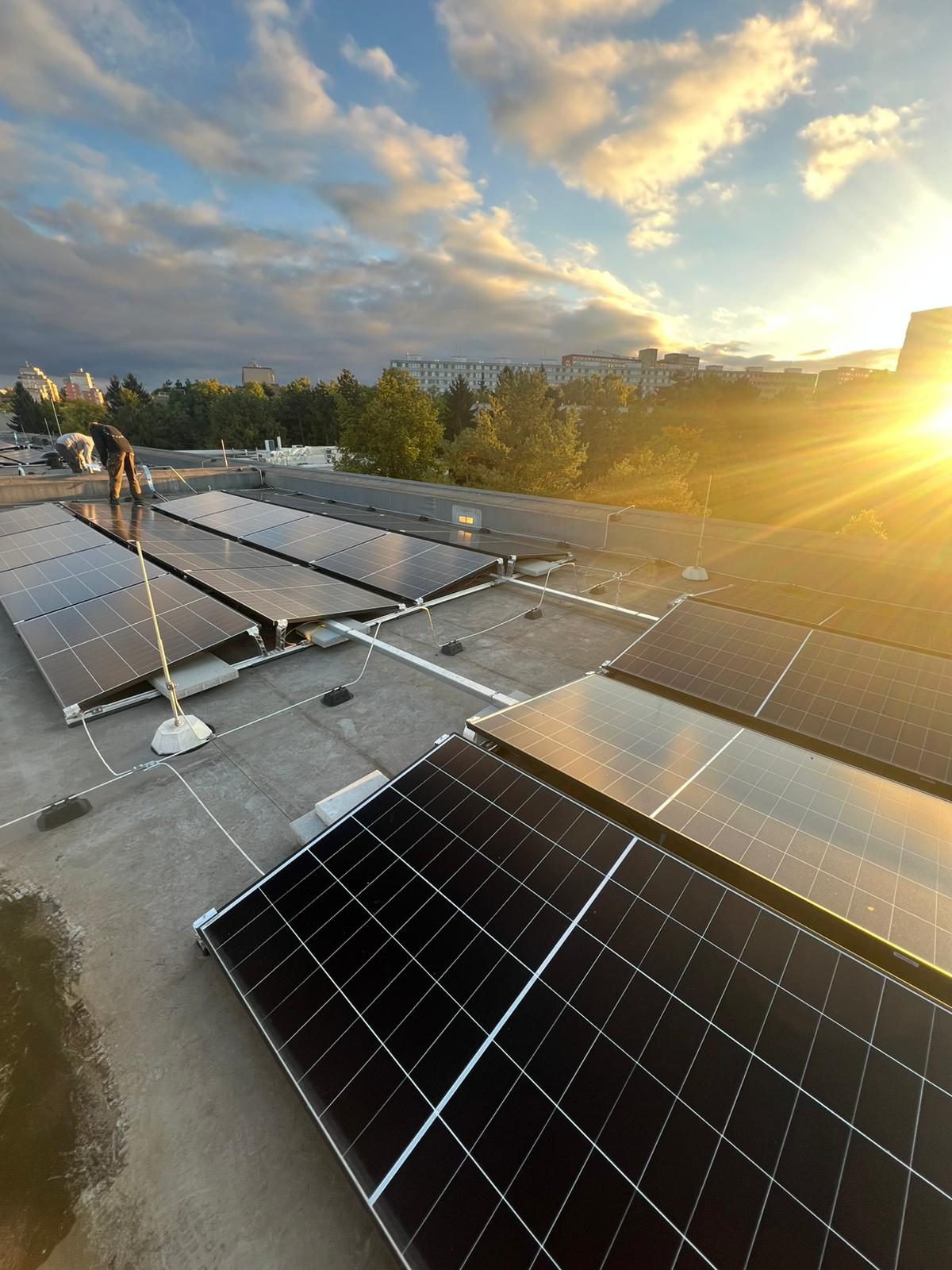Zlatavý podvečer na ploché komerční střeše s řadami černých fotovoltaických panelů a montérem v pozadí.