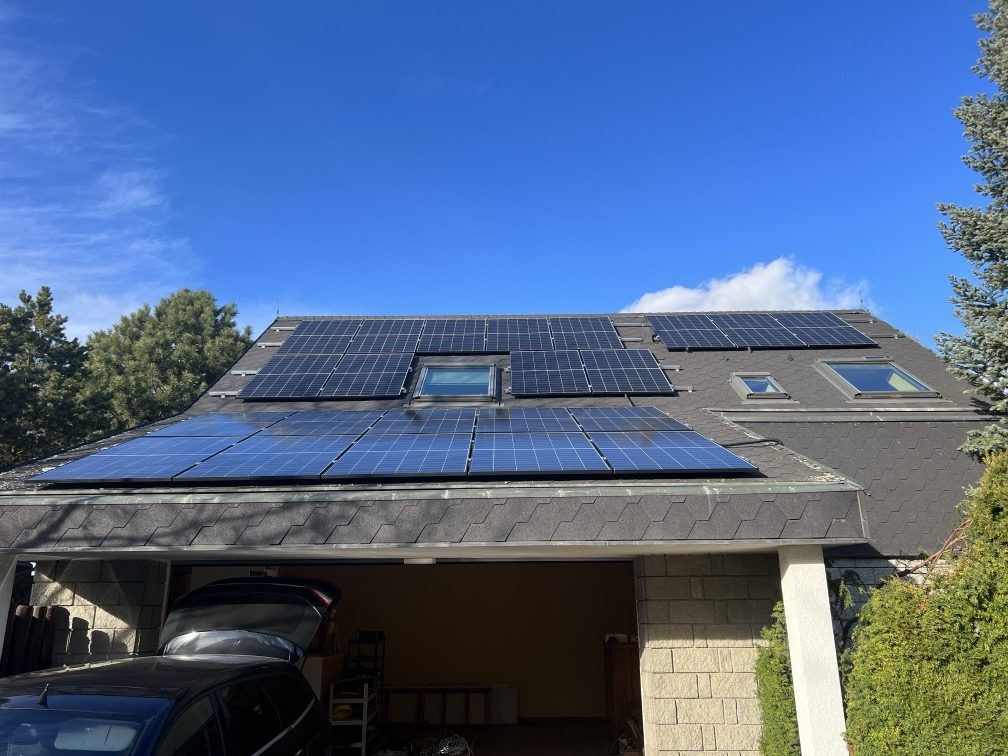 Modré fotovoltaické panely na tmavé střeše rodinného domu s garáží pod jasně modrou oblohou, rozmístěné kolem střešního okna, nedaleko Prahy.