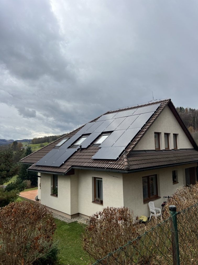 Rodinný dům na Valašsku s černými fotovoltaickými panely na hnědé sedlové střeše, v pozadí zalesněné kopce pod zataženou oblohou.
