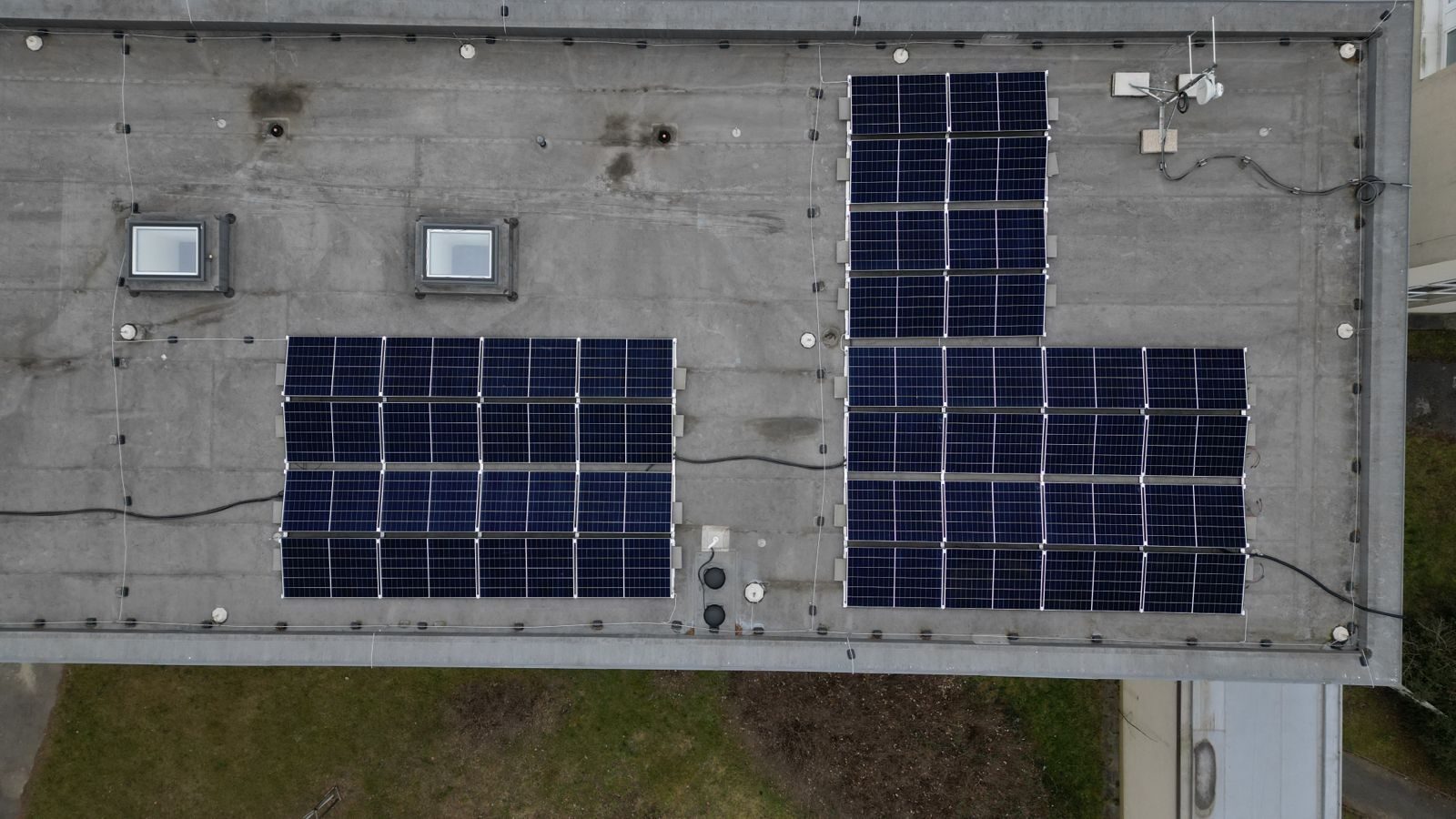 Pohled shora z dronu na plochou střechu komerční budovy v Praze s několika bloky tmavě modrých fotovoltaických panelů na šedé střešní ploše.